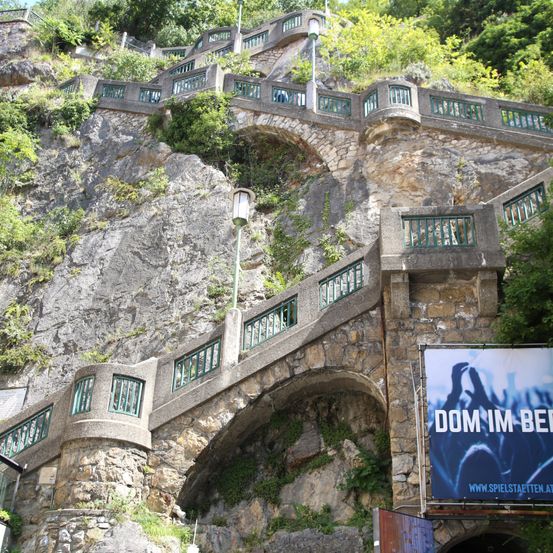 Eine Steintreppe mit grünen Geländern erklimmt einen steilen Felshang. Grüne Pflanzen wachsen am Felsen. Eine Straßenlaterne steht rechts. Ein Schild ist am Felsen angebracht.