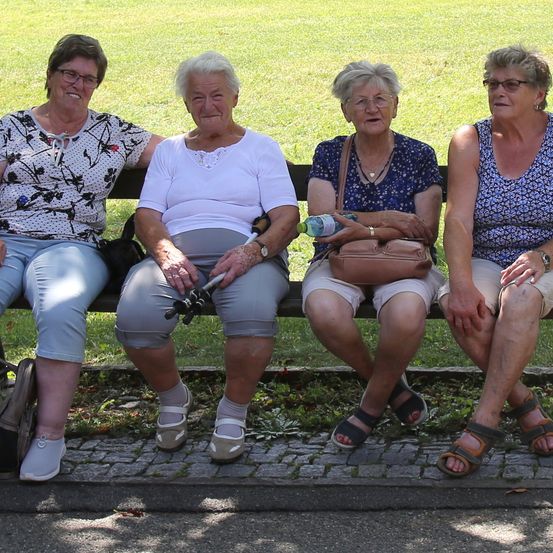Vier ältere Frauen sitzen auf einer Parkbank. Sie tragen alle eine Brille und Sandalen. Die Frau auf der linken Seite trägt ein Blumenhemd. Die mittlere Frau hat einen Stock.
