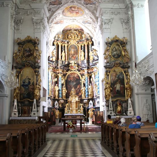 Ein Kircheninneres mit kunstvollen Altären und goldverzierten Statuen. Ein Tisch mit Blumen steht in der Mitte, und Menschen sitzen auf Bänken.
