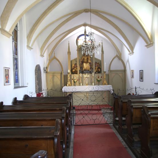 Innenansicht einer Kirche mit hölzernen Kirchenbänken, einem roten Teppich zum Altar und einem Altar mit Statue, Kerzen und Blumen.