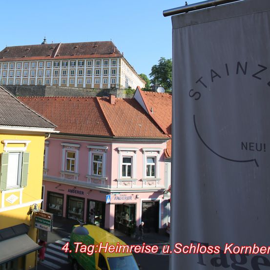 Ein Blick auf die Straße mit Gebäuden und einem gelben Lieferwagen davor. Die Gebäude sind in verschiedenen Farben gestrichen. Ein Banner hängt an der Straßenseite.