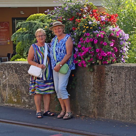 Zwei Frauen stehen neben einer Betonmauer mit Blumen. Eine trägt einen Hut und eine Uhr, die andere hat eine weiße Handtasche. Sie lächeln und scheinen für ein Foto zu posieren. Hinter ihnen steht ein Gebäude mit einem Schild und Grünpflanzen.