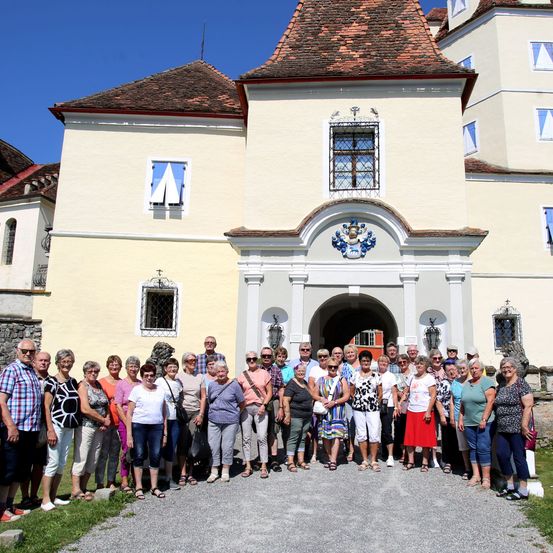 Eine Gruppe von Menschen posiert für ein Foto vor einem alten, historischen Gebäude mit mehreren Fenstern und einem Wappen über dem Eingang.