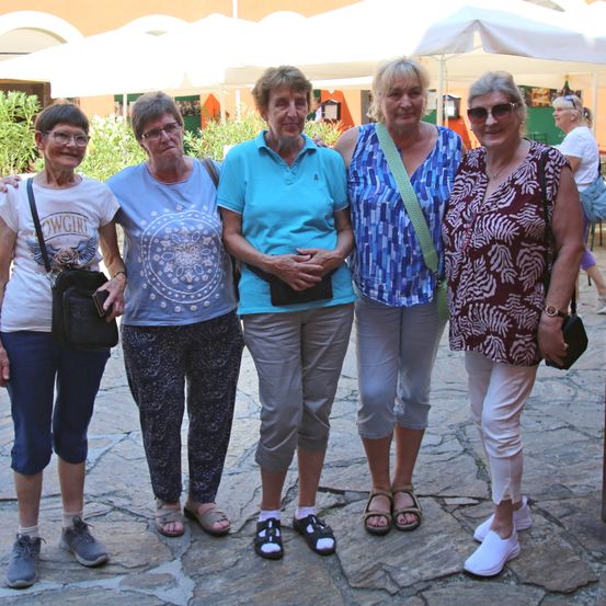 Fünf Frauen stehen lächelnd draußen, mit weißen Schirmen im Hintergrund. Sie tragen Freizeitkleidung und scheinen Touristen zu sein.