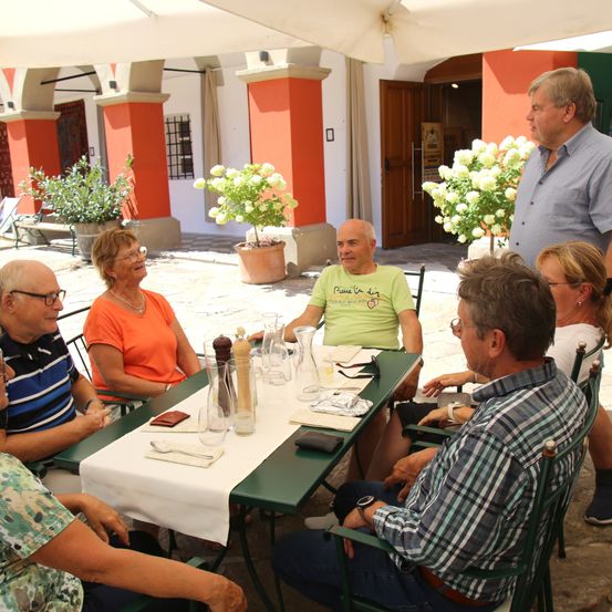 Eine Gruppe älterer Menschen sitzt in einem Außenrestaurant um einen Tisch herum, lächelt und führt ein Gespräch. Der Tisch ist mit Gläsern, Tellern und einer Vase gedeckt. Der Mann, der in der Nähe steht, spricht zu ihnen.
