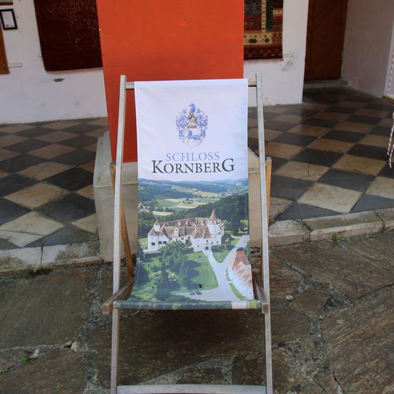 Ein äußerer Holzstuhl zeigt ein Banner mit einem Schlossbild und dem Namen Schloss Kornberg. Die Umgebung ist draußen, mit einem gefliesten Boden und einer offenen Tür.
