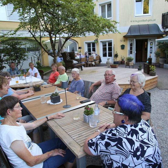 Eine Gruppe älterer Erwachsener sitzt um einen Tisch in einem Außenbereich, möglicherweise in einem Hotel, mit einem Gebäude im Hintergrund.
