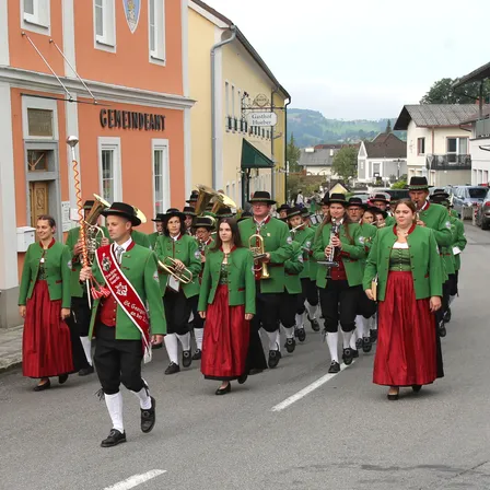 Eine Blaskapelle in grün und rot zieht in einem Umzug die Straße entlang, mit Gebäuden im Hintergrund.