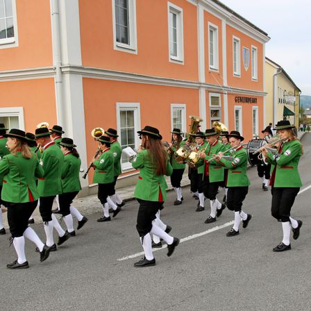 Eine Musikkapelle in grünen Uniformen spielt Musikinstrumente auf der Straße. Sie gehen in einer Reihe. Gebäude befinden sich am Straßenrand.
