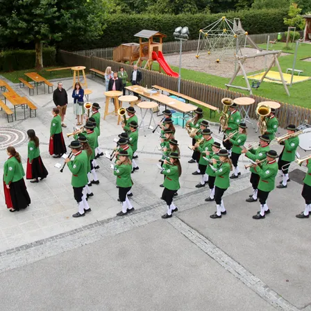 Ein Freiluftbereich zeigt eine Marschkapelle in grünen Uniformen, die mit verschiedenen Blechblasinstrumenten auftritt, mit Bänken, einer Rutsche und einem Spielplatz im Hintergrund.