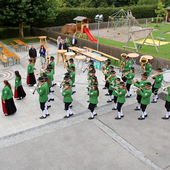 Ein Freiluftbereich zeigt eine Marschkapelle in grünen Uniformen, die mit verschiedenen Blechblasinstrumenten auftritt, mit Bänken, einer Rutsche und einem Spielplatz im Hintergrund.