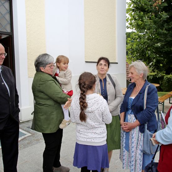 Eine Gruppe von Menschen steht vor einer Kirche, darunter eine ältere Frau, die ein kleines Kind hält. Andere Frauen, eine mit geflochtenen Haaren, führen ein Gespräch. Ein Mann und eine weitere Frau sind ebenfalls anwesend.
