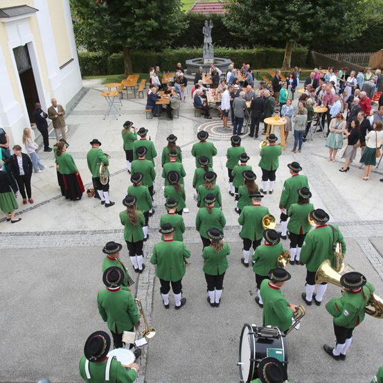 Eine Vogelperspektive einer traditionellen Blaskapelle in grünen Uniformen, die vor einem Gebäude mit einem Brunnen im Hintergrund aufführt.