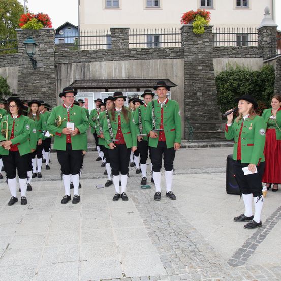 Eine Gruppe von Musikern in grünen Uniformen und schwarzen Hosen spielt vor einem Gebäude. Sie halten Musikinstrumente in der Hand und einer von ihnen singt in ein Mikrofon.