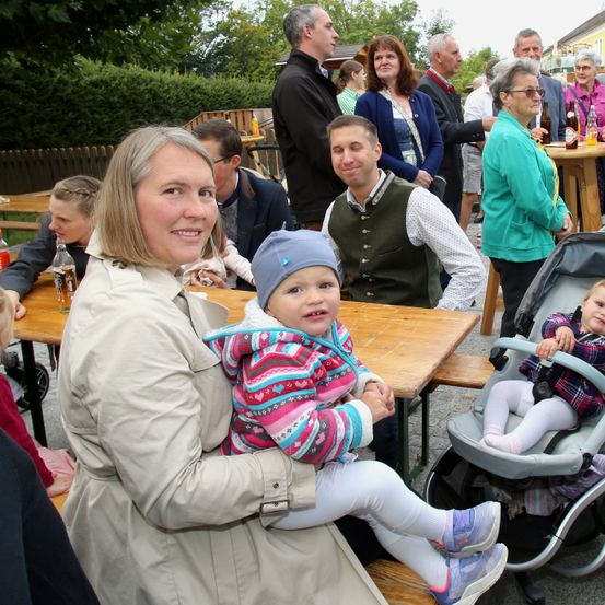 Eine Familie mit zwei kleinen Kindern, darunter ein Baby in einem Kinderwagen, sitzt an einem Picknicktisch. Eine Frau hält ein Kleinkind auf dem Schoß, während ein Mann hinter ihnen steht. Im Hintergrund versammeln sich Menschen an Tischen, einige mit Flaschen.