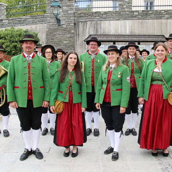 Eine Gruppe von Menschen in traditioneller grüner und roter Kleidung steht in einer Reihe vor einem Steinwall. Sie tragen Hüte und Schuhe, und einige halten Musikinstrumente. Im Hintergrund ist eine Steinmauer zu sehen.