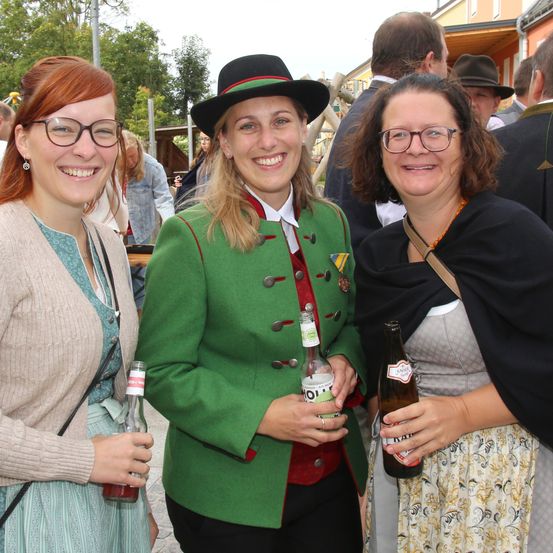 Drei Frauen in traditioneller Kleidung, lächelnd, halten Bierflaschen. Die Frau in der Mitte trägt eine grüne Jacke mit roten und schwarzen Akzenten. Der Hintergrund zeigt eine geschäftige Veranstaltung mit weiteren Menschen.