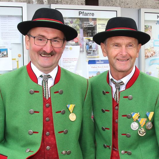 Zwei Männer in traditioneller deutscher Kleidung mit Medaillen um den Hals, lächeln in die Kamera, stehen nebeneinander. Hinter ihnen sind Poster mit Veranstaltungsinformationen sichtbar.