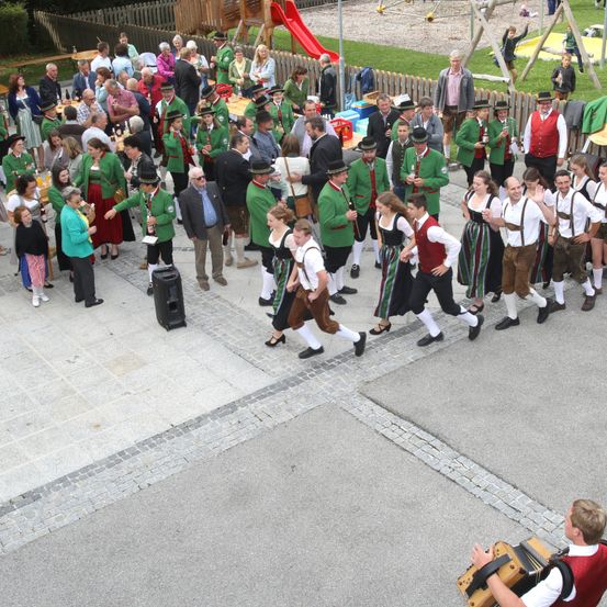Ein Outdoor-Event zeigt eine große Menschenmenge in grünen und weißen Kostümen, die tanzen und Instrumente spielen. Tische und Stühle sind aufgestellt, und ein Lautsprecher ist links. Ein roter Schlitten und ein Spielplatz im Hintergrund.