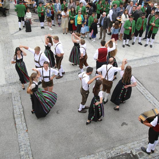 Eine Gruppe von Menschen tanzt in traditionellen Kostümen bei einer Veranstaltung im Freien. Sie sind von einer Menge von Zuschauern umgeben. Einige der Tänzer spielen Akkordeon.
