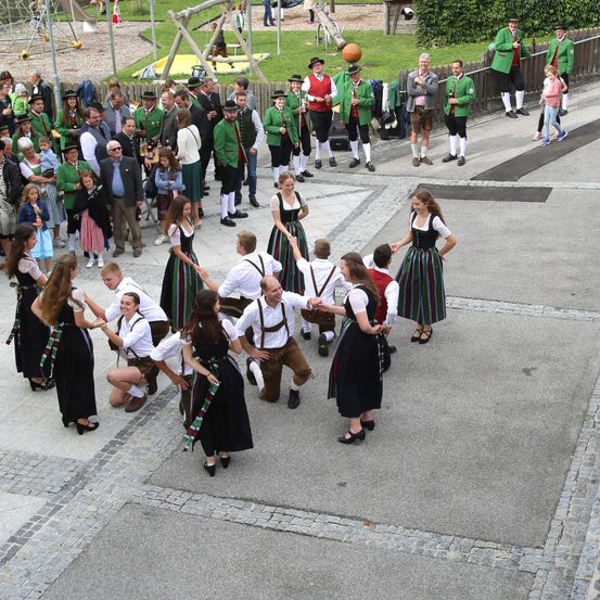 Eine Gruppe von Menschen in traditioneller Kleidung tanzt im Kreis. Einige tragen Hüte und Lederschuhe. Der Bereich ist mit Kopfsteinpflaster gepflastert. Hinter ihnen befindet sich ein Spielplatz.
