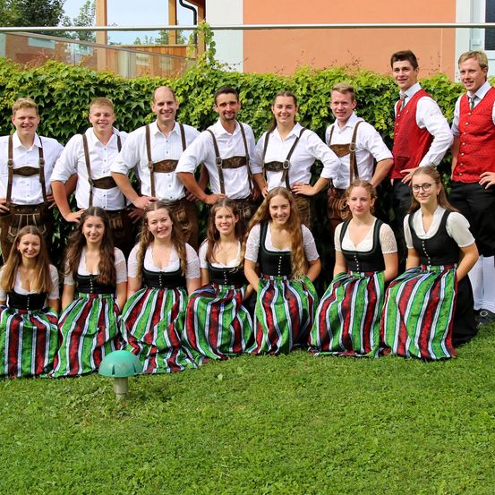 Eine Gruppe von Personen in traditioneller bayerischer Kleidung posiert für ein Foto auf einem grünen Rasen vor einem Gebäude.