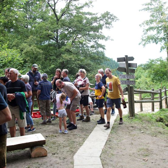 Eine Gruppe von Menschen steht im Freien, einige halten Flaschen. Ein Mann beugt sich vor, um einem Kind zu helfen. Ein Schild zeigt 'PLANKENSTEIN SCHIEBS'. Bäume und ein Holzzaun umgeben das Gebiet.