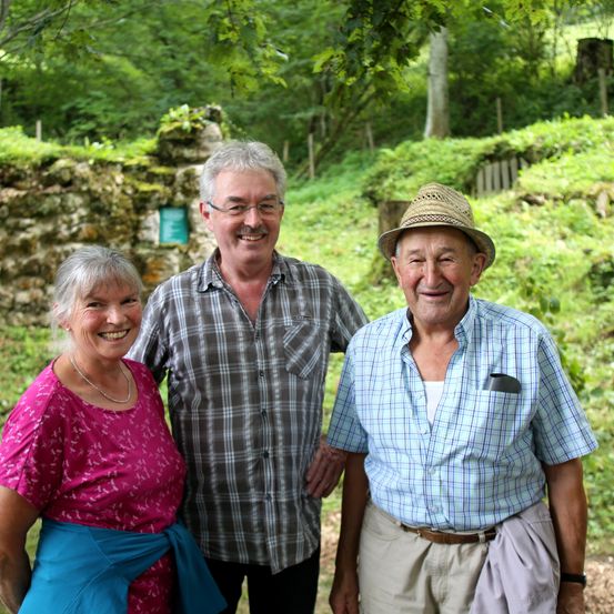 Drei Personen, eine Frau und zwei Männer, stehen gemeinsam in einem bewaldeten Bereich und lächeln für ein Foto.