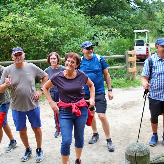 Eine Gruppe von Menschen läuft auf einem Pfad und posiert für ein Foto. Die Frau in der Mitte trägt ein lila T-Shirt und einen roten Schal. Dahinter befindet sich ein Holzzaun und ein Golfwagen.