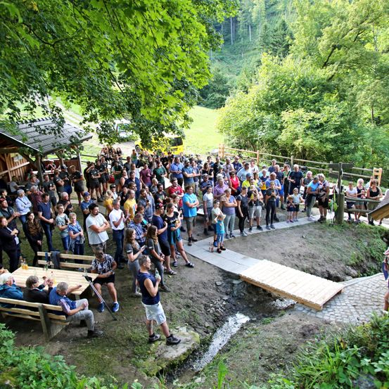 Eine Menschenmenge versammelt sich im Freien in einem bewaldeten Gebiet, einige sitzen auf Bänken, andere stehen in der Nähe einer Holzbrücke. Die Umgebung ist üppig mit Bäumen und Grün.