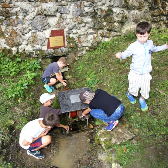 Vier Jungen erkunden einen kleinen Wasserbrunnen in der Nähe einer Steinmauer. Ein Junge hockt im Brunnen, ein anderer beugt sich hinunter und untersucht etwas. Ein dritter Junge kniet in der Nähe, und der vierte steht und beobachtet.
