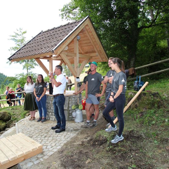 Eine Gruppe von Menschen steht um eine Holzhütte in einem Park. Ein Mann trinkt aus einer Flasche, während andere zusehen. Ein Zaun und Bäume sind im Hintergrund.