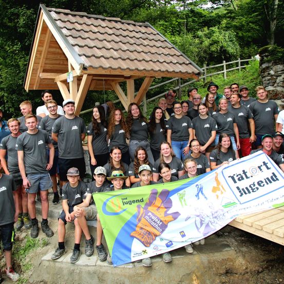 Eine Gruppe von Menschen in gleichen grauen T-Shirts posiert für ein Foto vor einer Holzstruktur und einem Banner mit Text in einer Fremdsprache.