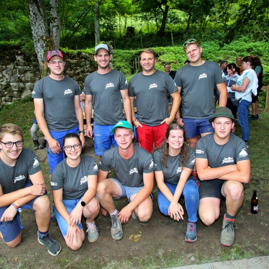 Eine Gruppe von Menschen in grauen T-Shirts mit Text und Logos posiert für ein Foto in einem Park. Sie lächeln und haben verschiedene Accessoires. Dahinter ist eine Steinmauer und Bäume zu sehen.