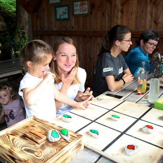Eine Frau und ein Mädchen spielen ein Spiel auf einem Holztisch. Das Mädchen hat ein Lächeln im Gesicht. Es gibt bemalte Steine auf dem Spielbrett. Drei andere Leute sitzen in der Nähe.