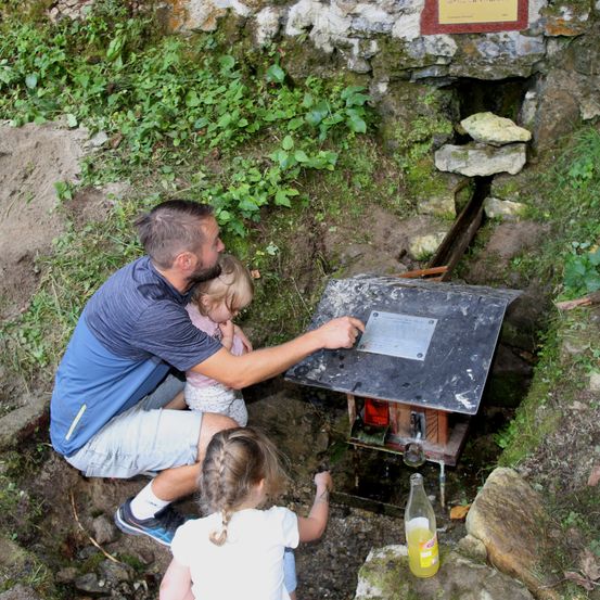 Ein Vater und zwei Töchter erkunden eine kleine Wasserpumpe in der Nähe einer moosbewachsenen Felswand. Das Mädchen rechts schaut sich die Pumpe an, während das Mädchen links sich am Vater lehnt.