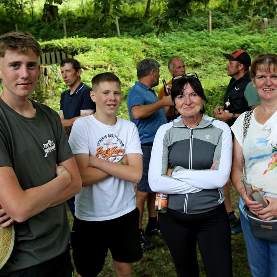 Eine Gruppe von Menschen steht im Freien. Zwei Frauen und zwei Jungen lächeln in die Kamera. Eine Frau hält eine Flasche und ein Mann in einem blauen Shirt hält eine Flasche in der Hand.