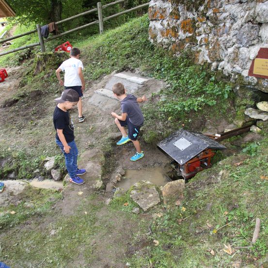 Drei Kinder erkunden einen schlammigen Bereich mit einer kleinen Struktur und einem Holzzaun. Ein Kind kniet in der Nähe der Struktur.
