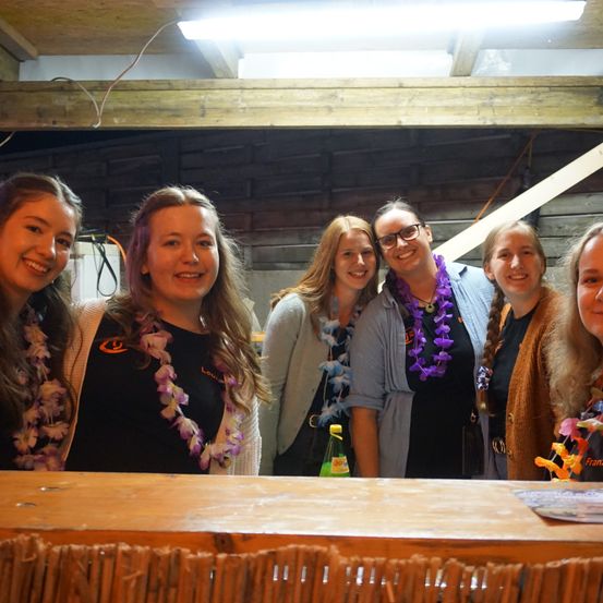 Eine Gruppe von sechs Frauen mit Kränzen posiert für ein Foto in einem rustikalen Ambiente, mit einer Holzbar und Ziegelwänden im Hintergrund.