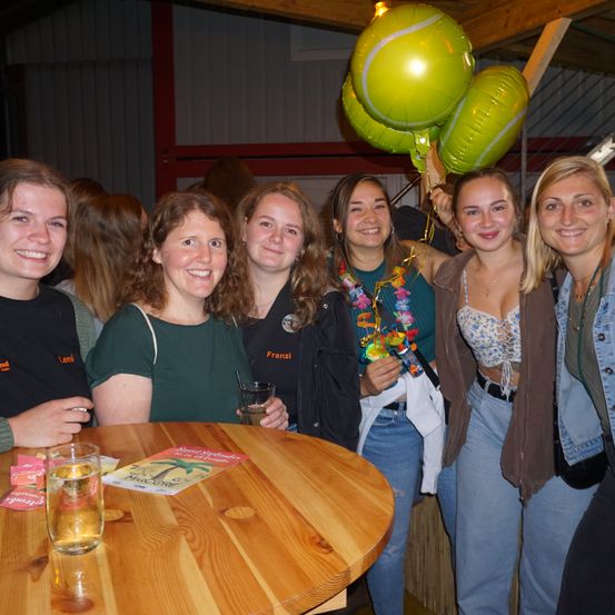 Eine Gruppe lächelnder Frauen, wahrscheinlich auf einer Party, mit grünen Tennisball-Luftballons im Hintergrund. Ein Tisch mit einem Buch und Getränken im Vordergrund.