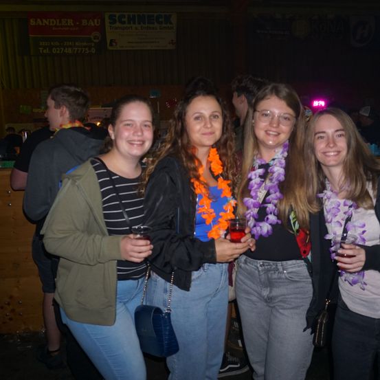 Vier Frauen stehen in einer Bar zusammen, lächeln und halten Getränke in den Händen. Sie tragen Blumenkränze um den Hals. Ein Banner hinter ihnen sagt 'Schneck' und eine Telefonnummer.