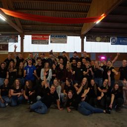 Eine Gruppe von Menschen in schwarzen T-Shirts posiert für ein Foto in einem Saal. Einige halten ihre Hände hoch, andere sitzen auf dem Boden. Werbung hängt an den Wänden.