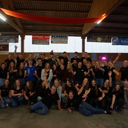 Eine Gruppe von Menschen in schwarzen T-Shirts posiert für ein Foto in einem Saal. Einige halten ihre Hände hoch, andere sitzen auf dem Boden. Werbung hängt an den Wänden.