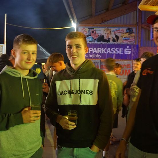 Zwei junge Männer in Kapuzenpullovern stehen bei Nacht in einem überfüllten Außenbereich, halten Getränke in den Händen und lächeln. Ein Banner hinter ihnen bewirbt PARKKASSE.