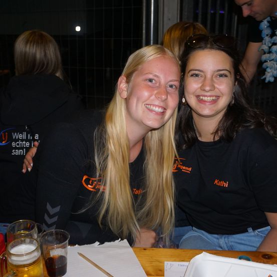 Zwei Frauen in schwarzen T-Shirts mit orangefarbenen Logos lächeln in die Kamera. Sie sitzen an einem Tisch mit Getränken, und andere sind im Hintergrund.