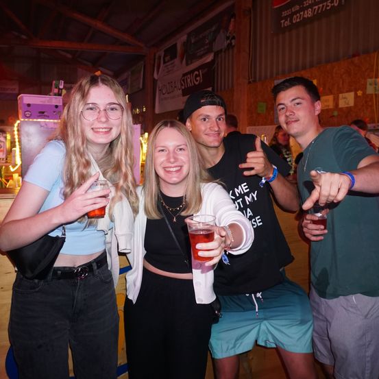 Vier junge Leute posieren für ein Foto bei einer nächtlichen Veranstaltung, zwei Frauen und zwei Männer, mit Getränken in der Hand, mit einer lächelnden Frau in der Mitte.