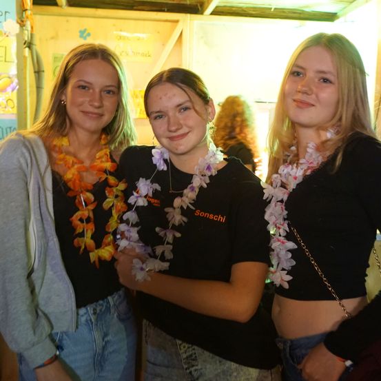 Drei Frauen mit Blumenketten stehen lächelnd zusammen. Eine trägt eine graue Kapuzenjacke, eine andere hat eine Blumenkette mit weißen Blumen und die dritte trägt ein schwarzes Top mit dem Wort 'Sonschi'.