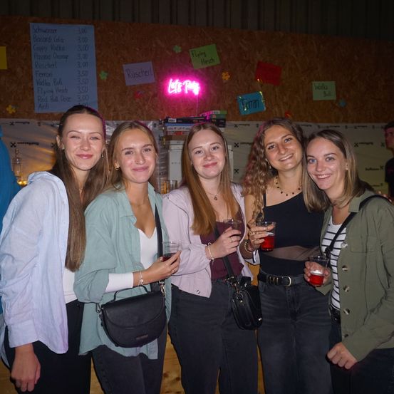 Fünf Frauen stehen lächelnd in einer Bar. Jede hält ein Getränk in der Hand, und ein Neonschild mit der Aufschrift 'Let's Party' ist hinter ihnen. Ein Menübrett mit verschiedenen Getränkeoptionen ist im Hintergrund zu sehen.