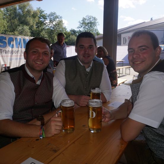 Drei Männer sitzen an einem Tisch, lächeln für die Kamera und halten jeweils ein Bierglas. Hinter ihnen steht ein Banner mit der Aufschrift 'SCHW'. Leute stehen in der Nähe von Tischen, und Bäume sind im Hintergrund zu sehen.