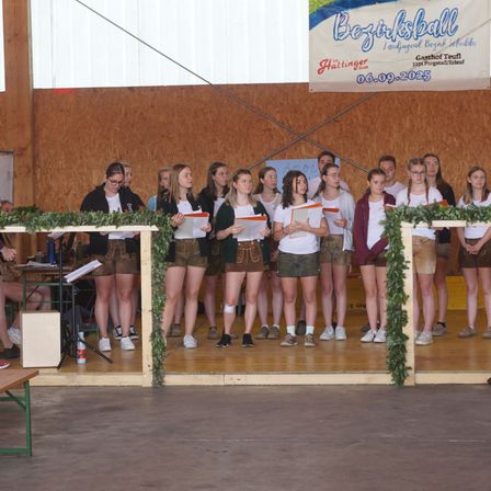 Eine Gruppe von Menschen steht auf der Bühne, hält Papiere in der Hand und trägt traditionelle Kleidung. Die Bühne ist mit Grünpflanzen dekoriert und hat ein Banner im Hintergrund.